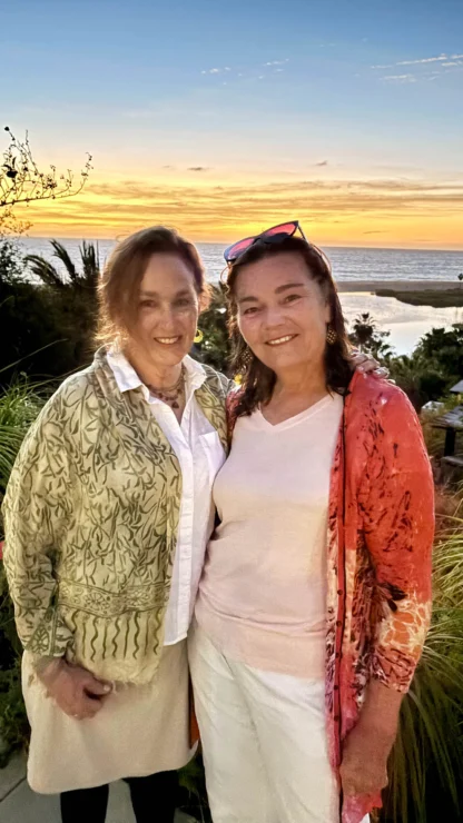 Two women smiling and standing together, with a scenic ocean sunset in the background. One wears a patterned jacket, and the other a coral cardigan.