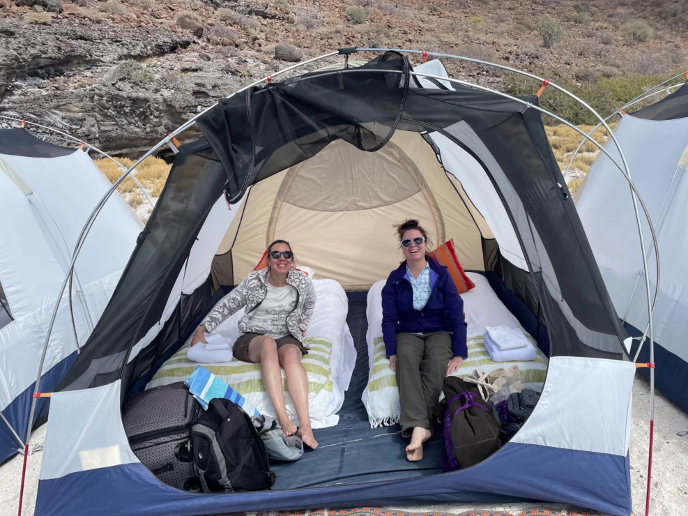 Two people relaxing in a tent with twin air mattresses and luggage, set in a rocky outdoor setting.
