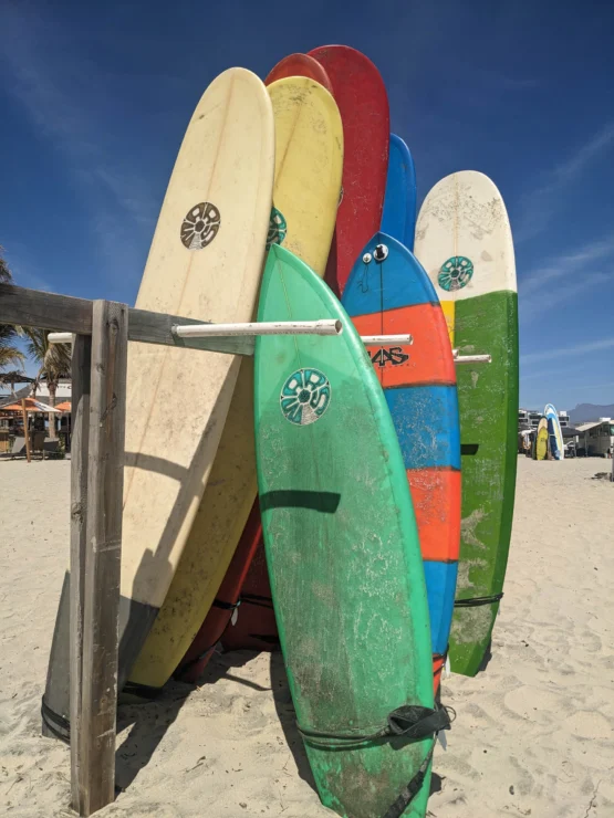 Seven colorful surfboards are standing upright on a sandy beach, leaning against a wooden rack, under a clear blue sky.