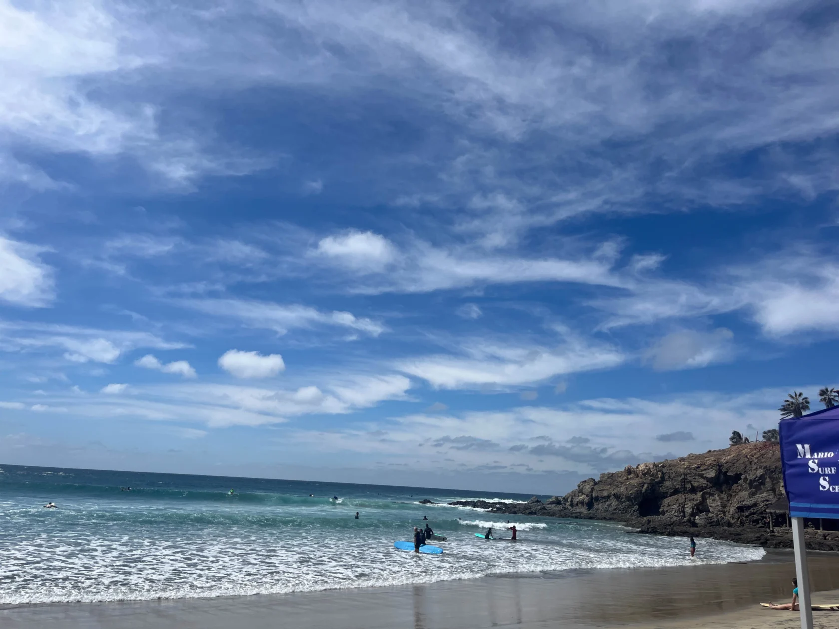 A beach scene with a blue sky, scattered clouds, and people in the ocean near a rocky shoreline. A blue sign is partially visible on the right side.