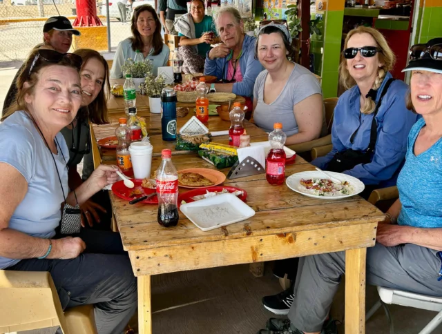 A group of people sitting around a wooden table with food and drinks in a casual outdoor setting.