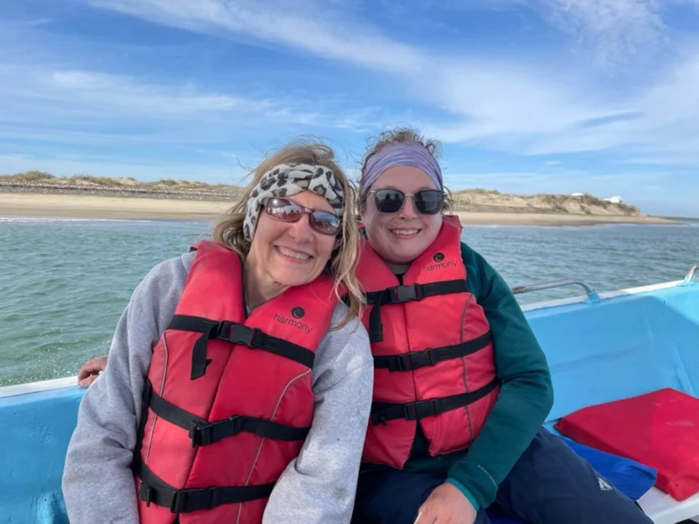 Two people wearing sunglasses and red life jackets sit on a boat with a coastline in the background.