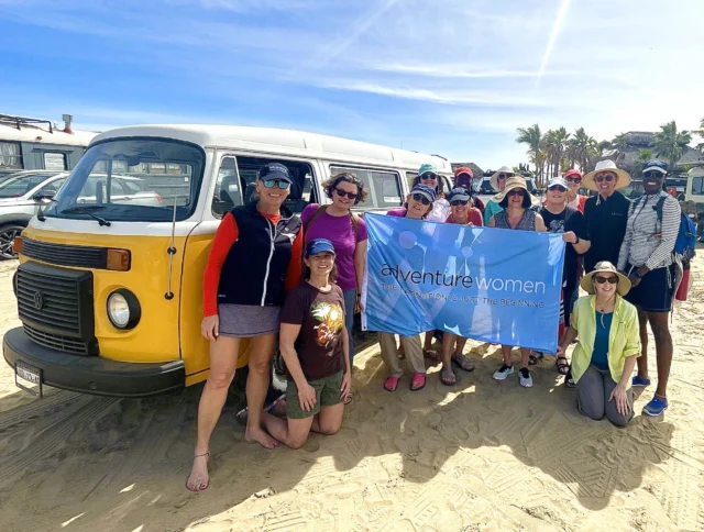A group of women standing on a beach, holding a banner that reads "Adventure Women." A yellow camper van is parked behind them.