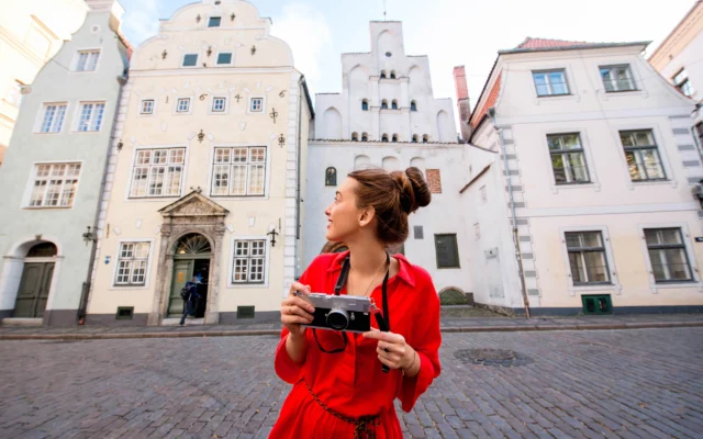 A person in a red outfit holds a camera, standing on a cobblestone street, surrounded by historic buildings with ornate facades.