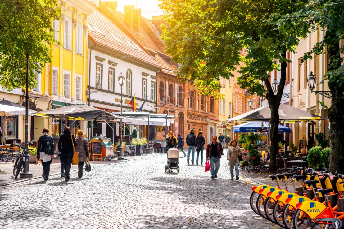 A cobblestone street with people walking, lined with vibrant buildings and outdoor cafes. Bicycles are parked in a row on the right. Sunlight filters through trees.