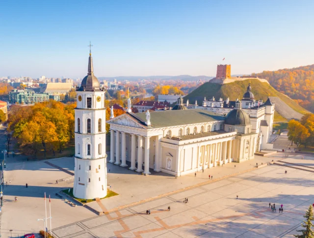Aerial view of a historic European city square with a white cathedral, bell tower, and hilltop castle. Autumn trees surround the area.