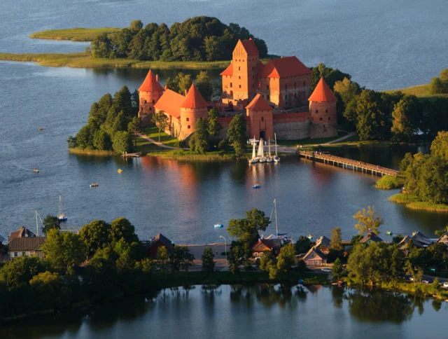 Aerial view of Trakai Island Castle, surrounded by a lake and connected to the mainland by a footbridge, with trees and boats nearby.