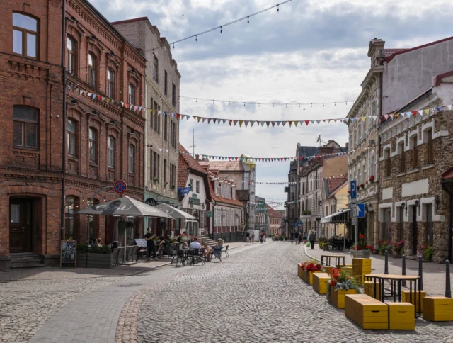A cobblestone street in a historic town with brick buildings, outdoor café seating, and colorful bunting hanging overhead.