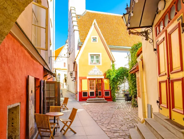 Narrow cobblestone street lined with colorful buildings; red, yellow, and pink facades. Wooden tables and chairs are set outside under a blue sky.