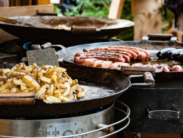Large pans cooking sliced potatoes and assorted sausages in an outdoor kitchen setting.