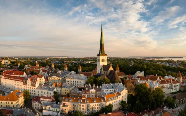 Aerial view of a historic cityscape with a tall church spire, surrounded by red-roofed buildings and greenery under a partly cloudy sky.