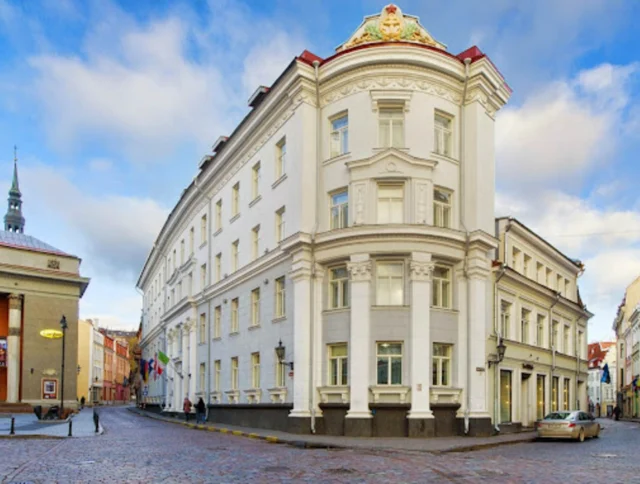 A large white historic building stands at a street corner with cobblestone roads, under a partly cloudy sky.