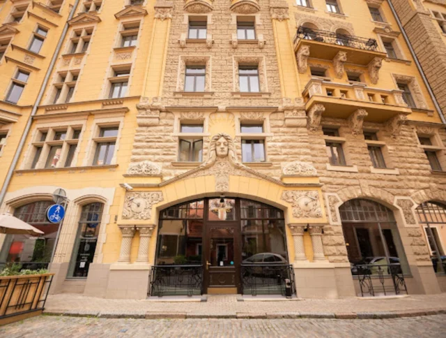 Facade of an ornate yellow building with intricate stonework, three arched entrances, and windows. A bicycle lane sign is visible on the left. Cobblestone street in front.