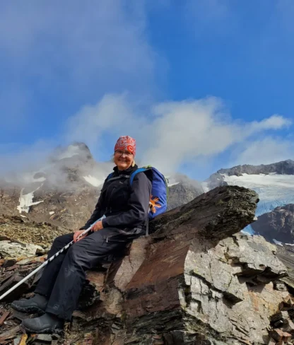 A person wearing hiking gear and a red bandana sits on a rock with a blue backpack, surrounded by rocky terrain and snow-capped mountains under a blue sky.