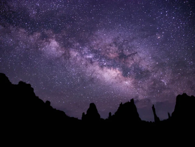 Night sky with a visible Milky Way galaxy, above silhouettes of rock formations.