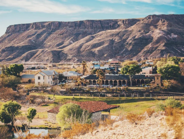 A small desert town with scattered houses, greenery, and a mountainous backdrop under a blue sky.