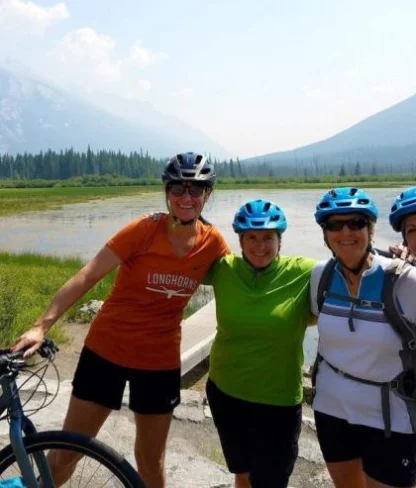 Four people wearing helmets stand with bicycles by a lake, surrounded by mountains and trees on a clear day.