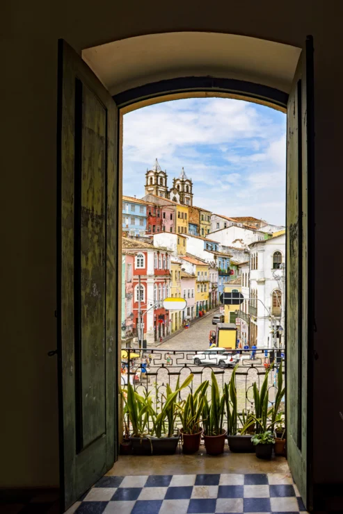 View through an arched doorway of a colorful historic street with a church in the background. Several potted plants line the doorway on a checkered floor.