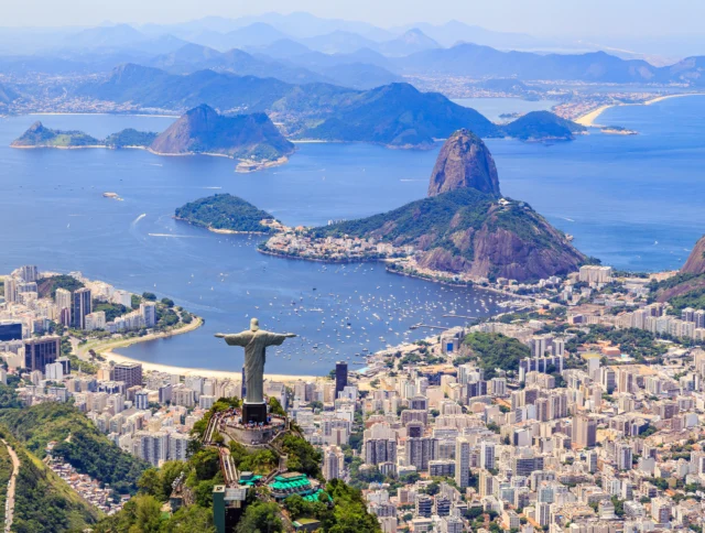 Aerial view of Rio de Janeiro, featuring the Christ the Redeemer statue, Sugarloaf Mountain, and the cityscape with surrounding ocean and mountains in the background.