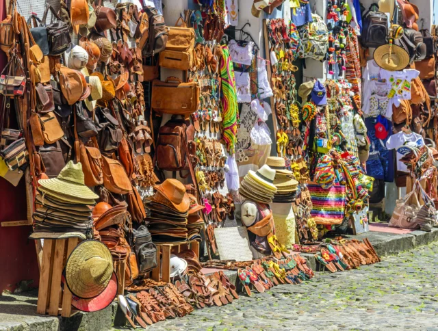 A market stall displays leather bags, hats, colorful textiles, and souvenirs on a cobblestone street.