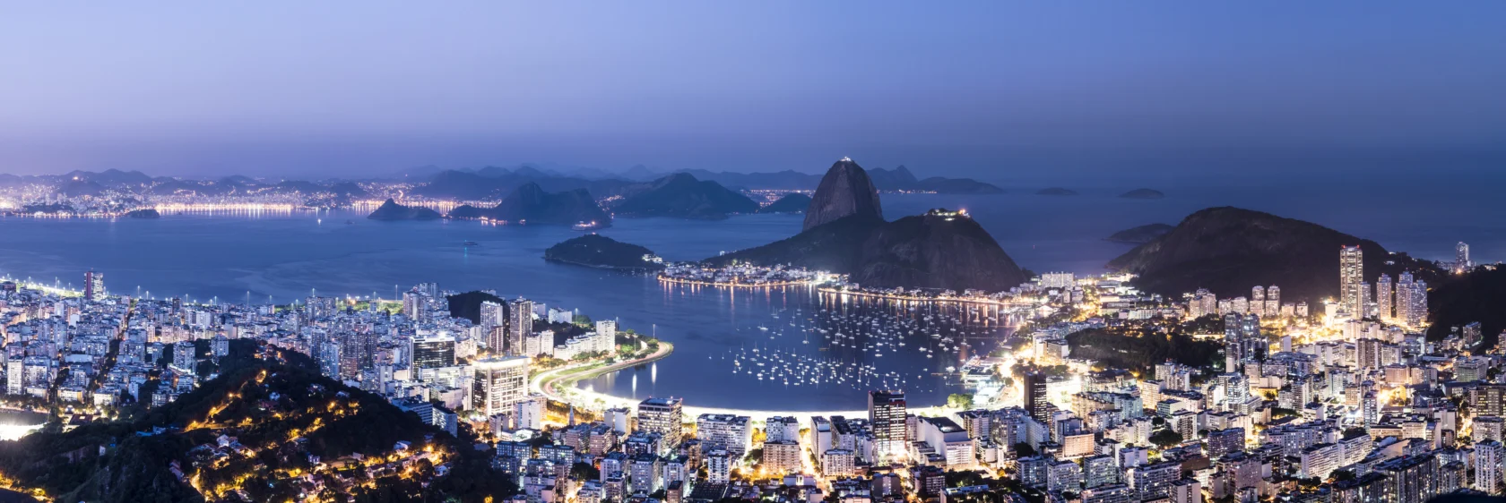 Panoramic view of a coastal cityscape at dusk with numerous buildings, a calm bay, and distant hills. Lights from the city illuminate the scene.