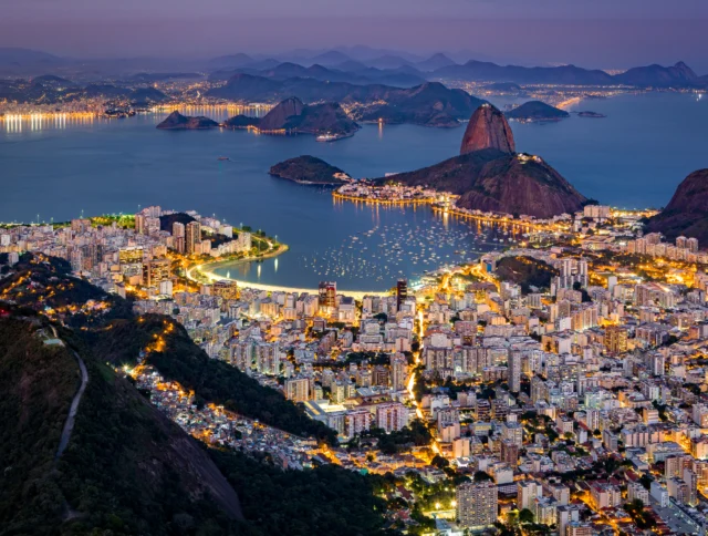 Aerial view of Rio de Janeiro at night, featuring illuminated cityscape, mountains, and ocean with boats.