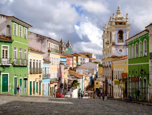 A cobblestone street lined with colorful colonial buildings under a partly cloudy sky, featuring a prominent church tower on the right. People are walking along the street.