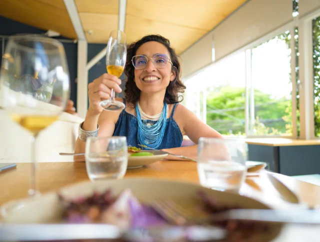 Woman sitting at a table in a restaurant, holding up a glass of wine and smiling. Plates and glasses are on the table in the foreground.