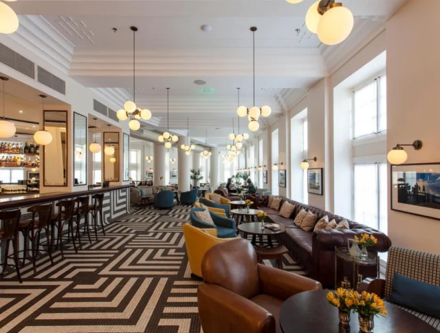 Spacious lounge area with patterned tile floor, cushioned leather chairs, and hanging globe lights. A bar is on the left, and the space has large windows.