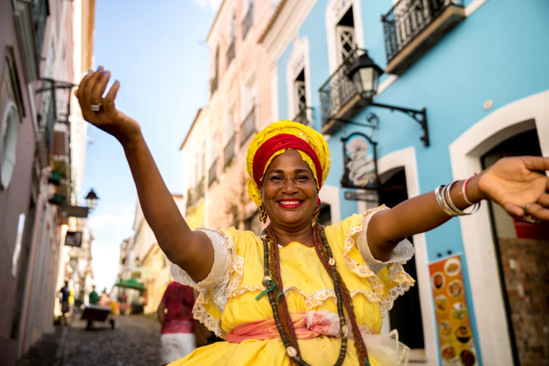 A woman in traditional dress smiles with arms raised on a colorful street with colonial buildings.