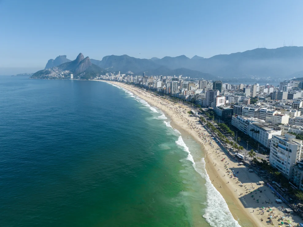Aerial view of a long beach in Rio de Janeiro, with waves along the shore, city buildings on the right, and mountains in the background under a clear blue sky.