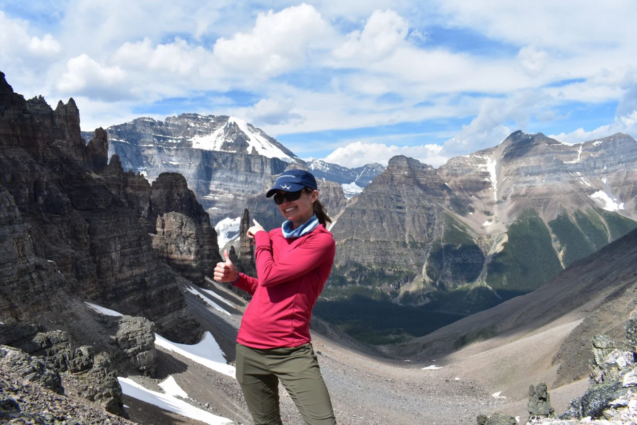 A person in a red shirt poses with a thumbs-up on a rocky mountain landscape under a blue sky with clouds. Snow-capped peaks are visible in the background.