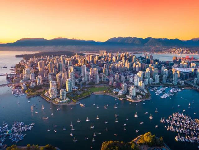 Aerial view of a city skyline at sunset with boats in the harbor, mountains in the background, and an orange sky.