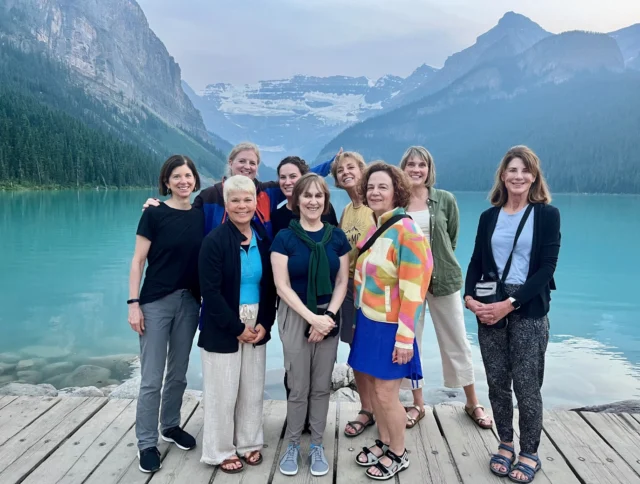 A group of nine women standing on a wooden dock in front of a turquoise lake, with mountains in the background.