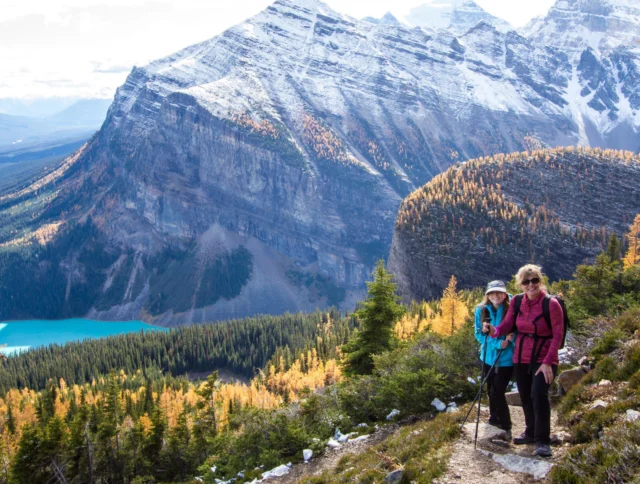 Two hikers walking on a mountain trail with snow-capped peaks and a turquoise lake in the background.