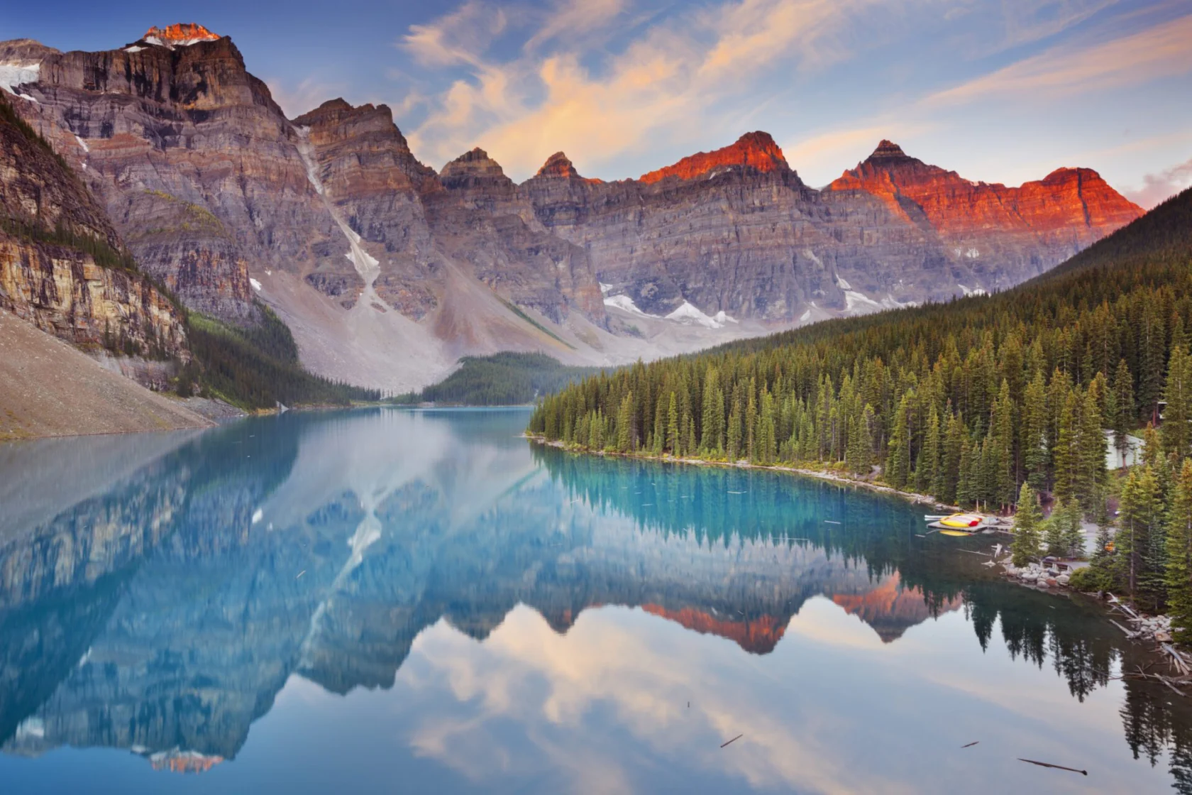 A serene lake reflects surrounding mountains and a forest under a partly cloudy sky at sunset.