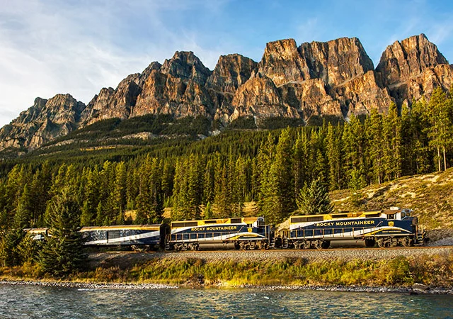 A train labeled "Rocky Mountaineer" travels alongside a river, with a backdrop of dense forest and rugged mountains under a blue sky.