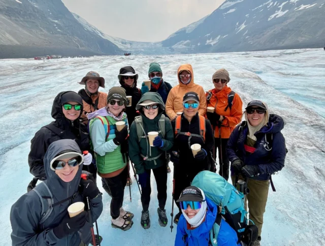 A group of people in outdoor clothing pose together on a glacier, holding cups, with snowy mountains in the background.