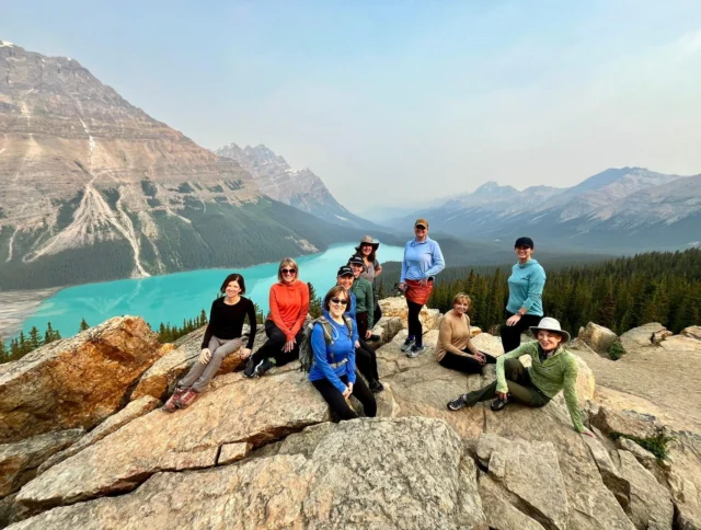 A group of people sitting on a rocky ledge overlooking a turquoise lake and mountains in the background.
