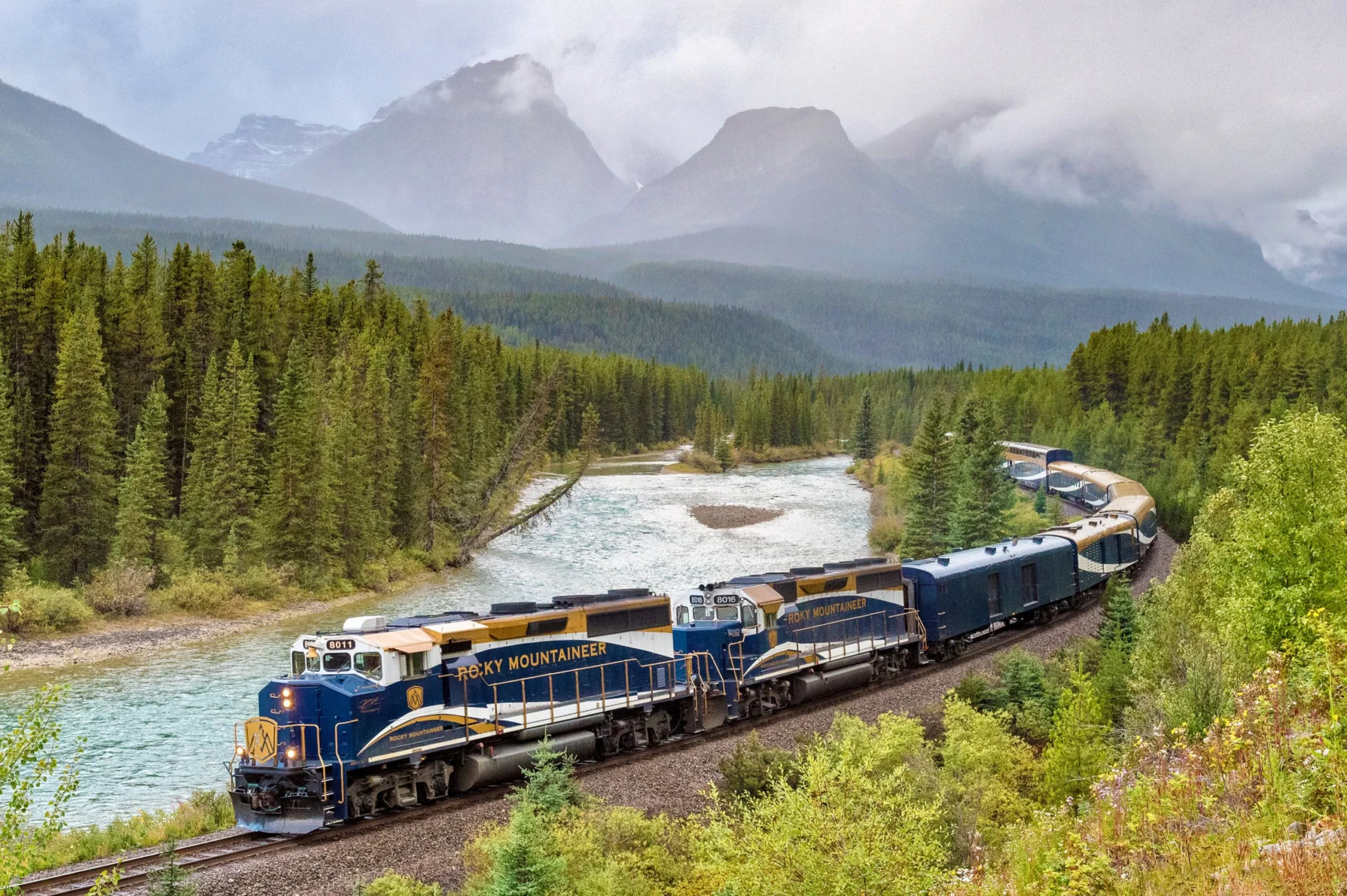 A blue and gold train travels through a forest alongside a river, with misty mountain peaks in the background.
