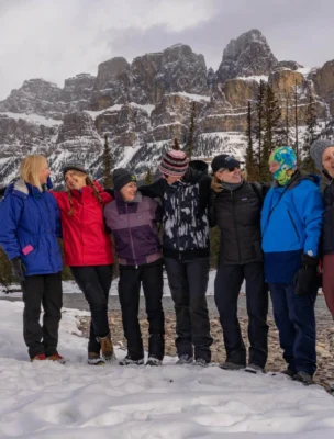 A group of people dressed in winter clothing stand together on snow-covered ground with a mountain range in the background.