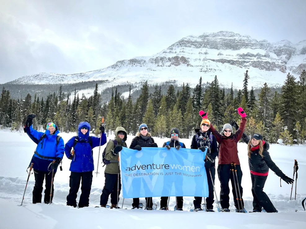 A group of women with hiking gear stand in the snow, holding a banner that reads "adventurewomen." Snowy trees and a mountain are in the background.