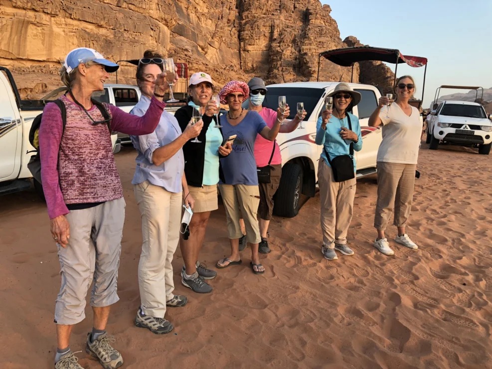 A group of people stands in a desert setting, holding up drinks and smiling, with rocky cliffs and parked vehicles in the background.