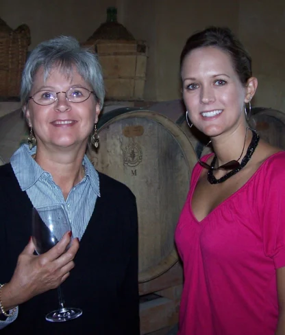 Two women standing in front of wine barrels. One holds a wine glass and wears glasses; the other wears a pink top.