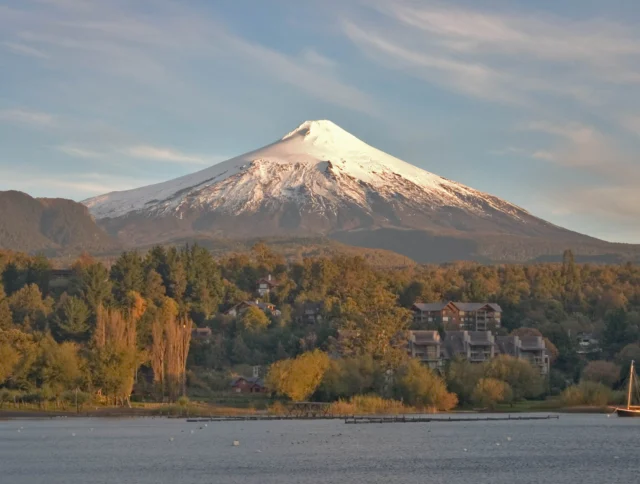 Snow-capped volcano with lush greenery and residential buildings in the foreground, under a clear blue sky.
