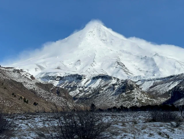 Snow-covered mountain peak with clear blue sky, surrounded by rocky terrain and sparse vegetation.
