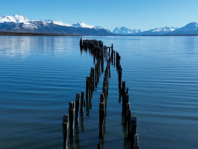 Old wooden piers extend into a calm blue lake with snowy mountains in the background under a clear sky.