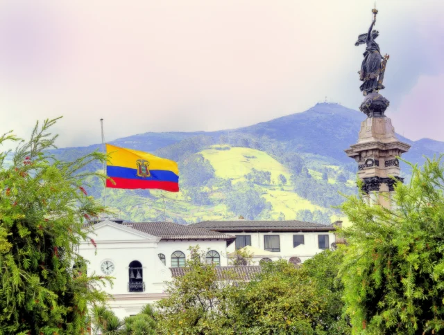 Ecuadorian flag next to a historic monument with a mountainous background and lush greenery in the foreground.