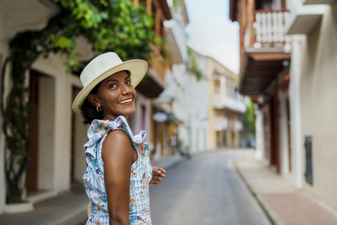 A woman in a floral dress and hat smiles while standing on a narrow street lined with colorful buildings and greenery.