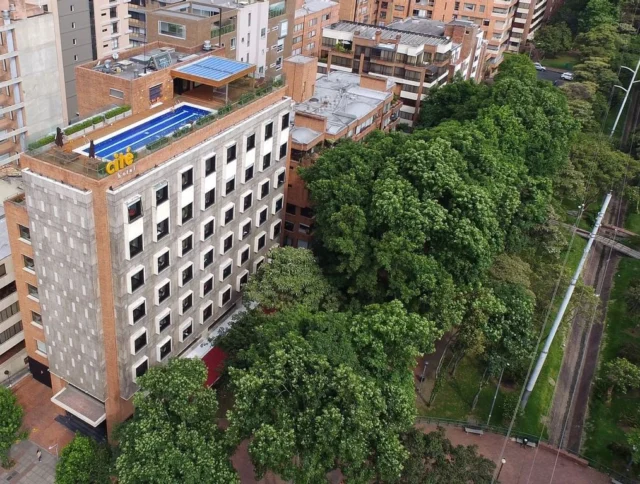 Aerial view of a multi-story building with a rooftop pool, surrounded by trees and adjacent to a road with light traffic.